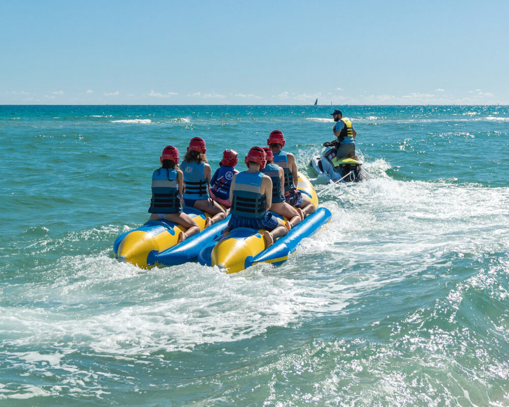 A group in red helmets rides a banana boat through turquoise surf behind a jet ski.