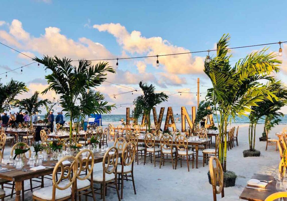 A long candlelit dinner table set on the sand at dusk.