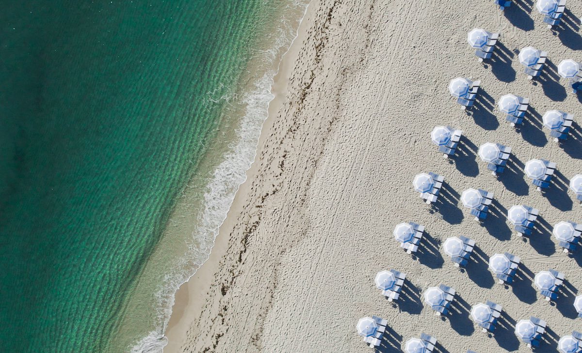 Aerial view of the Miami Beach shoreline: emerald water, white sand, and rows of Boucher Brothers umbrellas casting long morning shadows.